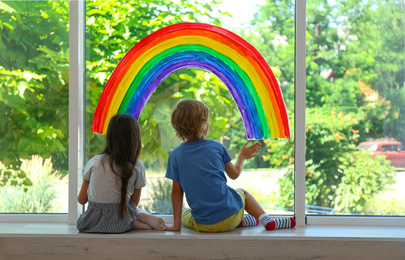 Little children sitting near rainbow painting on window indoors. Stay at home concept