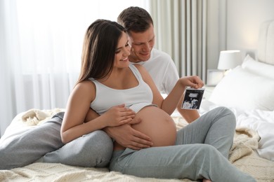 Photo of Young pregnant woman and her husband with ultrasound picture of baby in bedroom