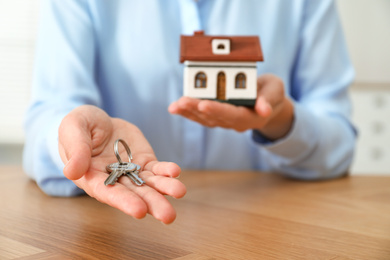 Real estate agent holding house model and keys at wooden table, closeup