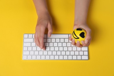 Woman squeezing antistress ball while typing on keyboard against yellow background, top view