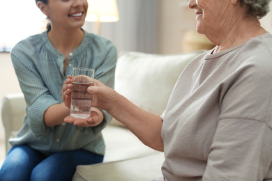 Young woman giving water to elderly lady indoors, closeup. Senior people care