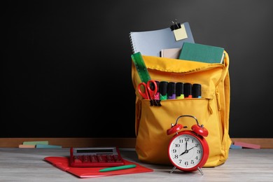 Backpack with different school stationery on white wooden table near blackboard