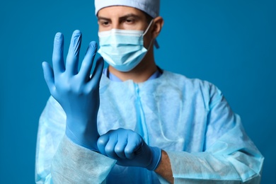 Doctor in protective mask putting on medical gloves against blue background