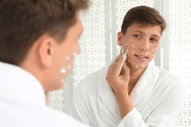 Teen guy with acne problem applying cream near mirror in bathroom