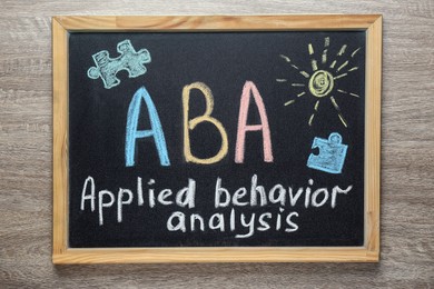 Photo of Small blackboard with text ABA Applied behavior analysis and drawings on wooden table, top view