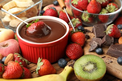 Photo of Fondue fork with strawberry in bowl of melted chocolate surrounded by other fruits on wooden table