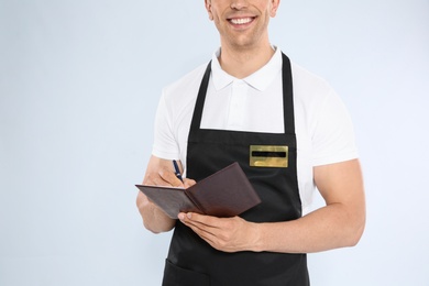 Young waiter in apron taking order on light background