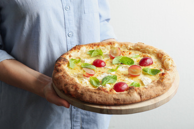 Photo of Woman holding delicious cheese pizza with grapes and basil on light background, closeup