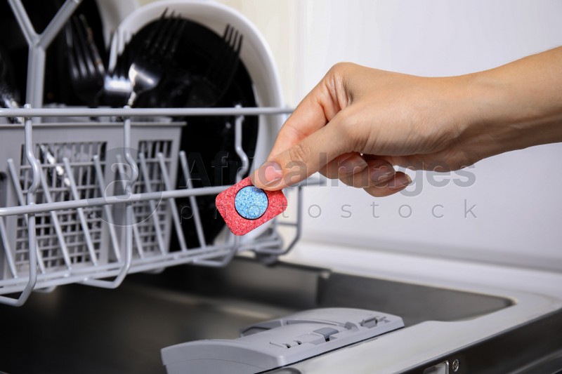 Photo of Woman putting detergent tablet into open dishwasher in kitchen, closeup
