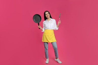 Young housewife with frying pan and spatula on pink background