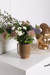 Stylish ceramic vase with beautiful flowers and golden Buddha sculpture on chest of drawers near white wall