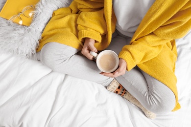 Woman with coffee wearing knitted socks on white fabric, top view. Warm clothes