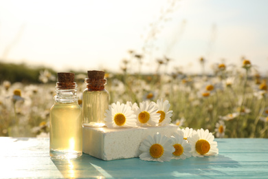 Bottles of essential oil, soap bar and fresh chamomiles on light blue wooden table in field