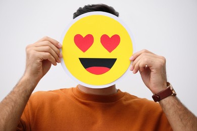 Man hiding emotions using card with drawn smiling face on white background