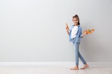 Little girl with brush and paint palette near light wall indoors. Space for text