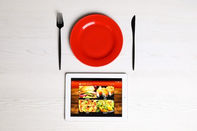Modern tablet with open page for online food ordering, plate and cutlery on white wooden table, flat lay. Concept of delivery service
