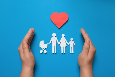 Man protecting paper family figures and red heart on light blue background, top view. Insurance concept