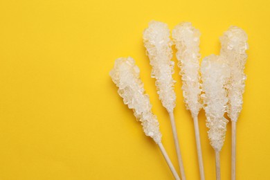 Wooden sticks with sugar crystals and space for text on yellow background, flat lay. Tasty rock candies