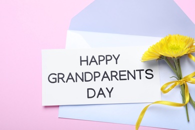 Card with phrase Happy Grandparents Day, envelope and beautiful flowers on pink background, flat lay