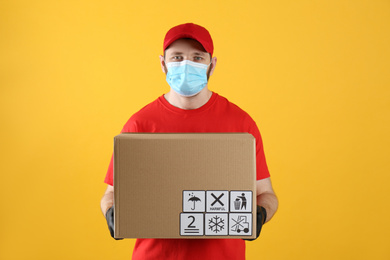 Courier in mask holding cardboard box with different packaging symbols on yellow background. Parcel delivery