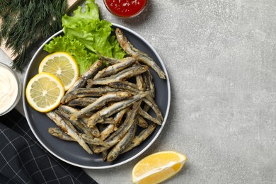 Photo of Delicious fried anchovies with lemon, lettuce leaves and sauces served on light grey table, flat lay. Space for text