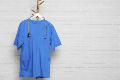 Photo of Blue medical uniform and stethoscope hanging on rack near white brick wall. Space for text