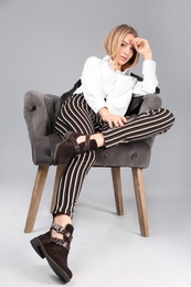 Young stylish woman with trendy shoes sitting in armchair on grey background