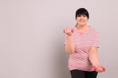 Happy overweight mature woman doing exercise with dumbbells on grey background, space for text