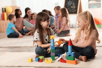 Cute girls playing with building blocks on floor while kindergarten teacher reading book to other children indoors