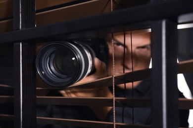 Private detective with camera spying near window indoors, focus on lens