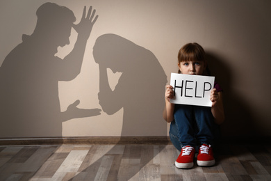 Sad little girl with sign HELP sitting on floor and silhouettes of arguing parents 