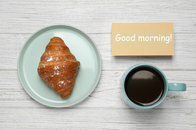 Photo of Croissant, coffee and card with words GOOD MORNING on white wooden table, flat lay. Tasty breakfast