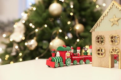 Bright toy train and decorative wooden house on white table near Christmas tree. Space for text