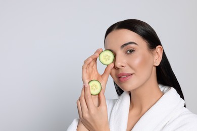 Photo of Woman in bathrobe holding pieces of cucumber on light grey background, space for text. Spa treatment