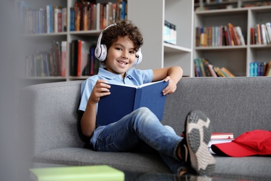Cute little boy in headphones reading book on sofa in library