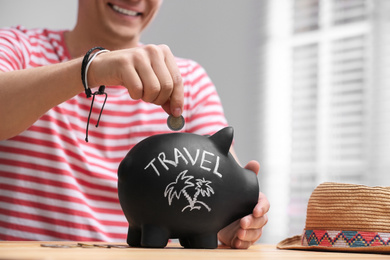 Man putting coin into piggy bank with word TRAVEL at table, closeup