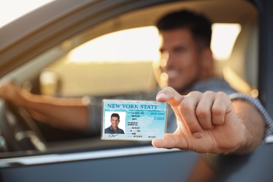 Happy man holding license while sitting in car outdoors, focus on hand. Driving school