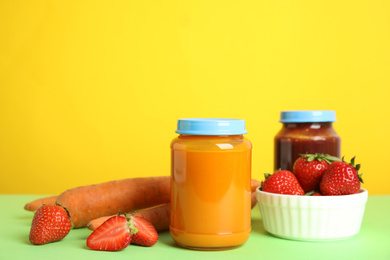 Jars with baby food and fresh ingredients on green table against yellow background