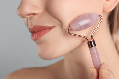 Photo of Young woman using natural rose quartz face roller on light grey background, closeup