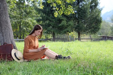Beautiful young woman drawing with pencil in notepad near tree on green grass