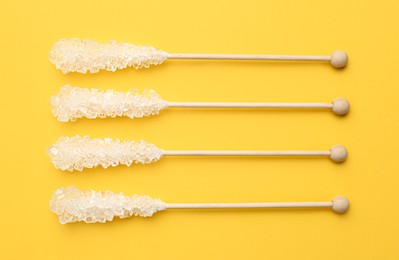 Wooden sticks with sugar crystals on yellow background, flat lay. Tasty rock candies