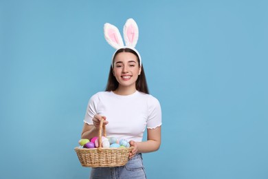 Photo of Happy woman in bunny ears headband holding wicker basket of painted Easter eggs on turquoise background