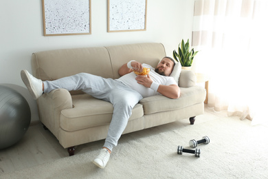 Lazy overweight man eating chips on sofa at home
