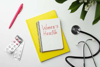 Notebook with text Women's Health, pills and stethoscope on white table, flat lay