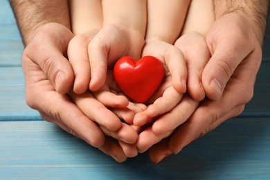 Parents and kid holding red heart in hands at light blue wooden table, closeup