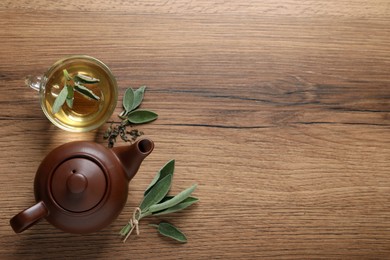 Cup of sage tea, green leaves and teapot on wooden table, flat lay. Space for text