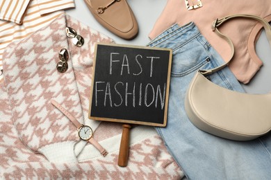 Small chalkboard with phrase FAST FASHION, stylish clothes and accessories on light background, flat lay