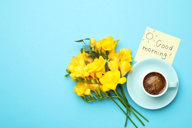 Cup of aromatic coffee, beautiful yellow freesias and Good Morning note on light blue background, flat lay. Space for text