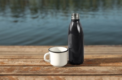 Modern black thermos bottle and cup on wooden surface near river