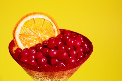 Photo of Martini glass with cranberries and dry orange slice on yellow background, closeup